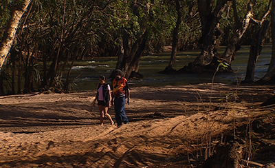 Two people walk along a shaded riverbank.