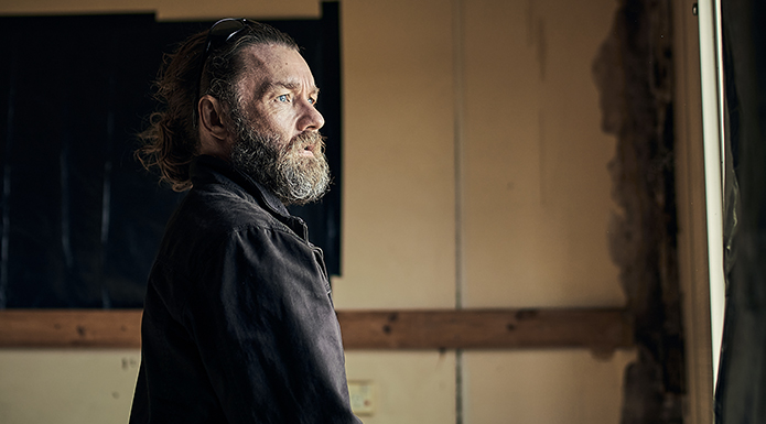 A photo of actor Joel Edgerton with shaggy hair and a beard, and sunglasses on his head, looking out a window from inside a decrepit room