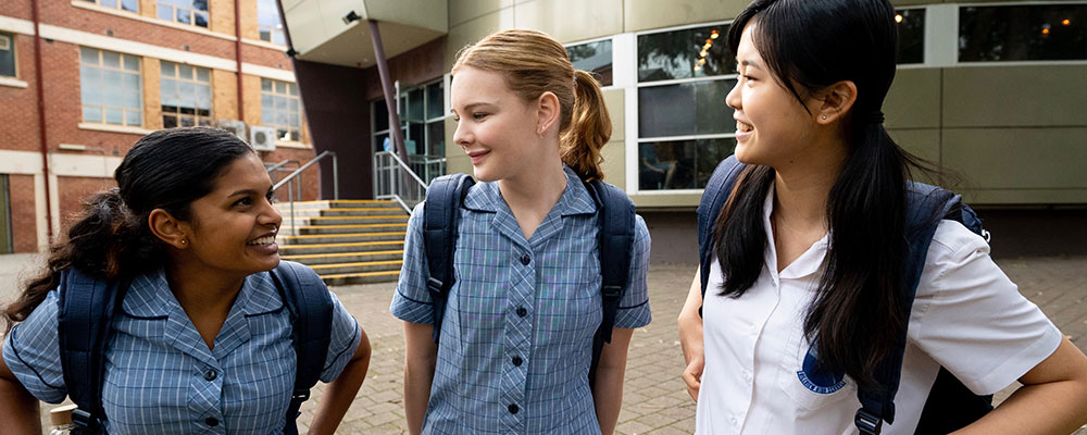 Three schoolgirls in uniform standing outside a school, smiling at each other