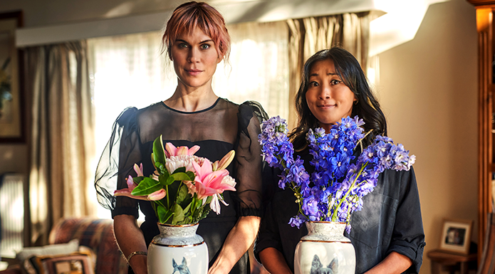 Two women stand side by side holding ceramic vases of flowers.