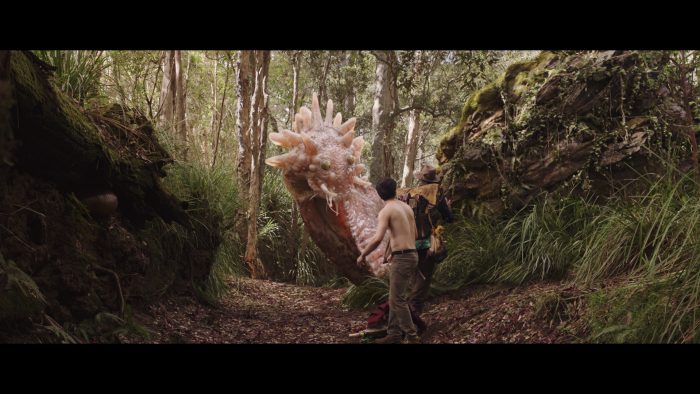 A still from Love and Monsters -a shirtless man in a forest looks at a slug-like pink monster