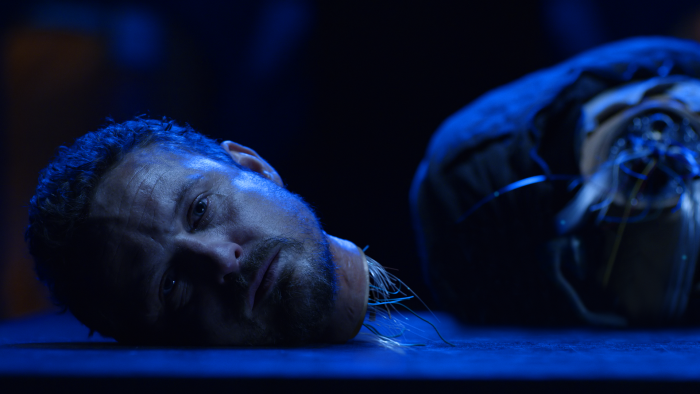 A mans head with robotic wires coming from the neck lies on a table