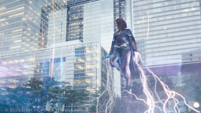 A woman wearing a superhero costume floats in the air in front of city building, with lightning pouring from her hands and feet