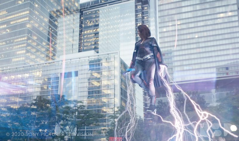 A woman wearing a superhero costume floats in the air in front of city building, with lightning pouring from her hands and feet