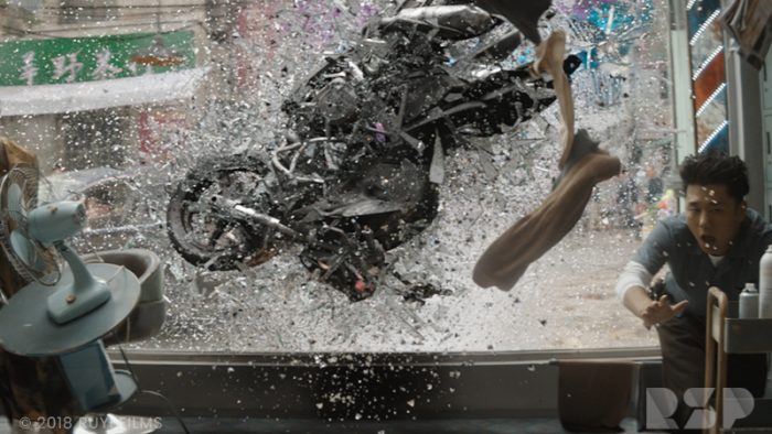 A motorbike smashes through a glass window, and a man wearing hospital scrubs gasps and jumps out of the way.