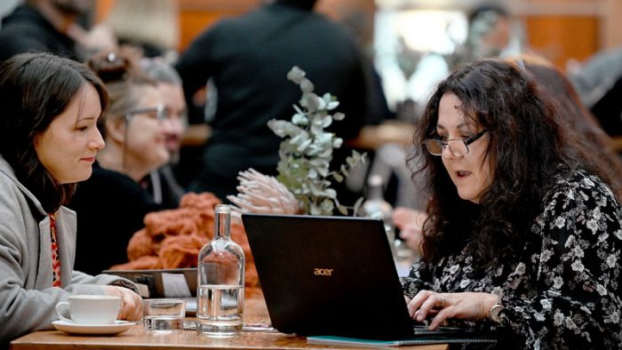 Two women with dark hair sit at a table looking into a laptop, a coffee and some water are placed nearby.