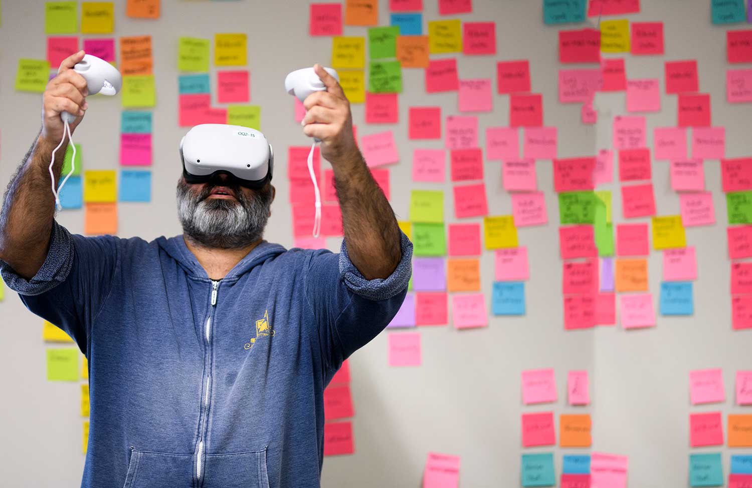 A man wearing a virtual reality headset and holding controllers standing in front of a whiteboard covered in colourful post-it notes