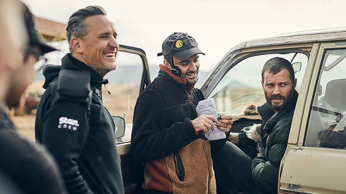 Two male crew members consulting on set with actor Jamie Dornan, who is seated inside a car