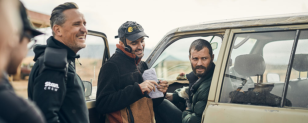 Two male crew members consulting on set with actor Jamie Dornan, who is seated inside a car