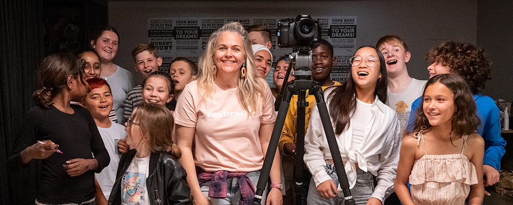 Taryn Brumfitt smiling and wearing a pink t shirt standing with a camera in front of a group of children smiling