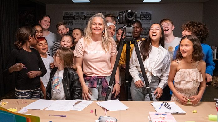 Taryn Brumfitt smiling and wearing a pink t shirt standing with a camera in front of a group of children smiling