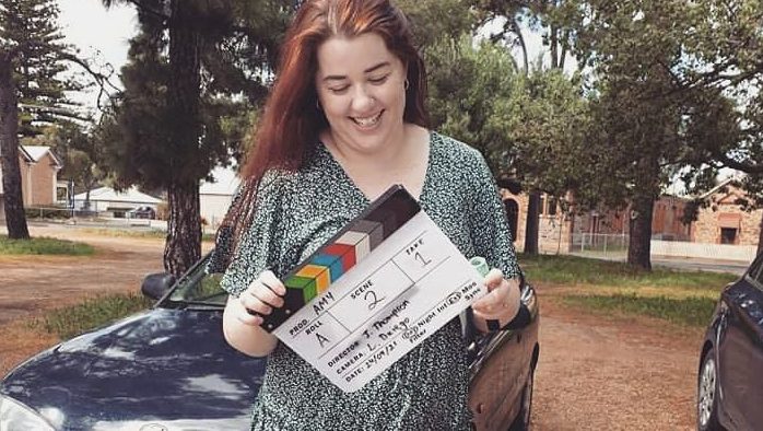 Star stands in front of a car and some trees holding a clapper board