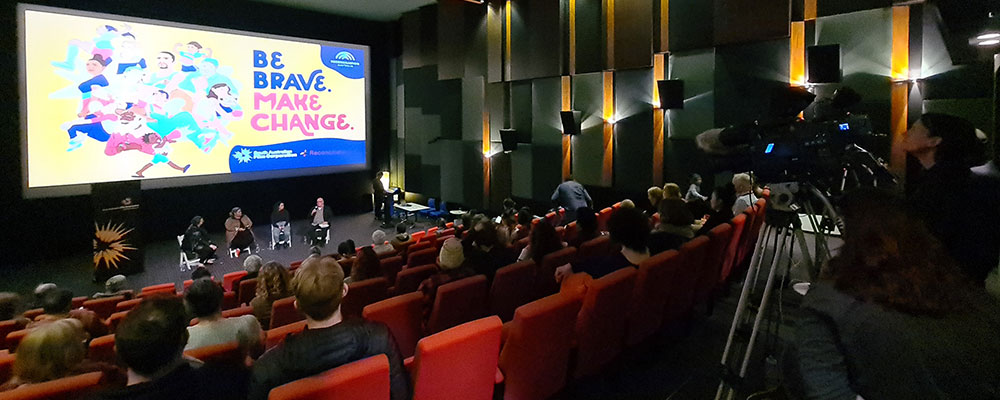 People sitting in a cinema watching four people speaking on stage, while a camera crew films them