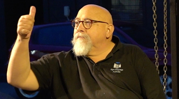 A man with a bald head, beard and glasses sits on a theatre stage giving the thumbs up