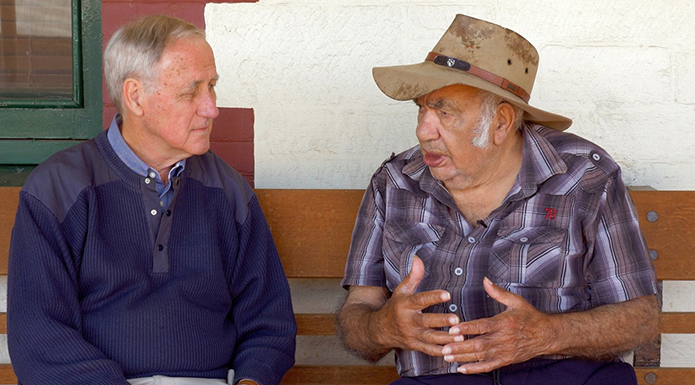 Two elderly men sit on a bench in conversation