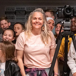 Taryn Brumfitt smiling and wearing a pink t shirt standing with a camera in front of a group of children smiling