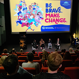 People sitting in a cinema looking at people on stage in front of a screen with words reading "Be Brave make change"