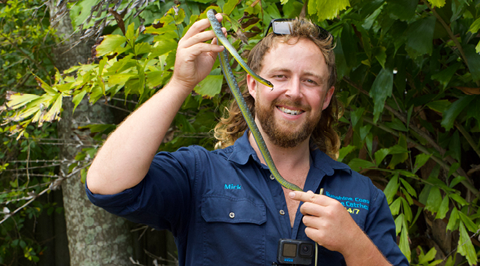 2025-TV-ASWDeadlyPursuit A man dressed in an Aussie Snake Wranglers shirt holds up a small green snake.