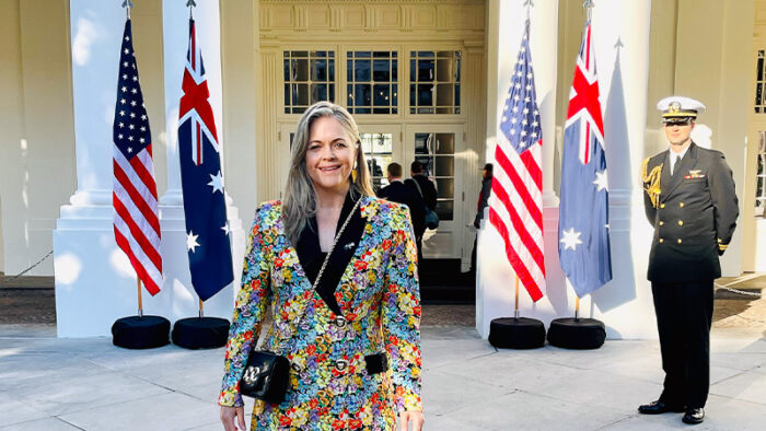 Taryn Brumfitt wears a colourful suit in front of a grand entrance with US and Australian flags displayed.
