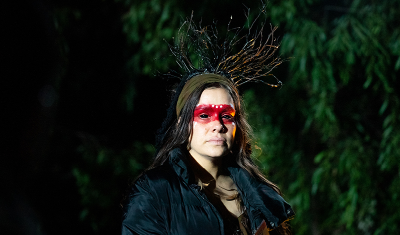 A woman wearing an elaborate headpiece that looks like glass branches stands in front of a tree at night. She has a band of red pain across her eyes, which are fully black.