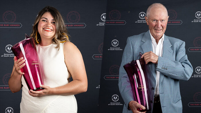 Two people stand in front of a media wall holding large red glass awards.