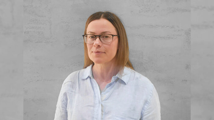 A woman in a light-coloured linen shirt stands in front of a grey background.