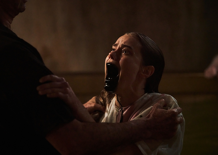 A young woman clutches a man who holds onto her as she screams. Black liquid pours out of her mouth.