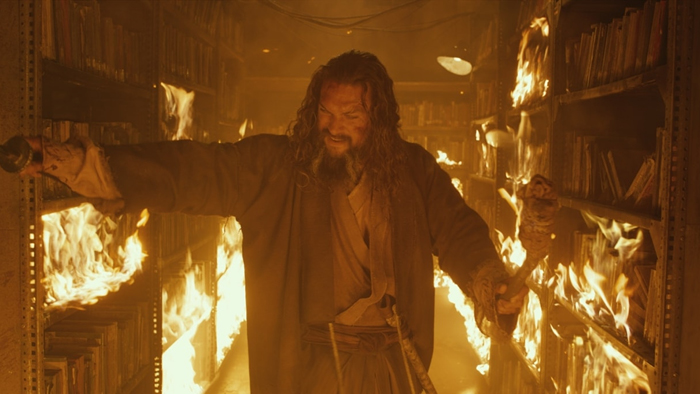 Jason Momoa in See stands among burning bookshelves.