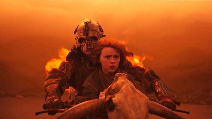 Young Furiosa sits on a flaming motorcycle in front of a rider in a skull helmet.