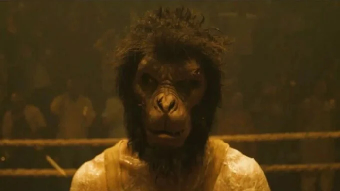 A man in a monkey mask stands in a wrestling ring, staring at the camera.