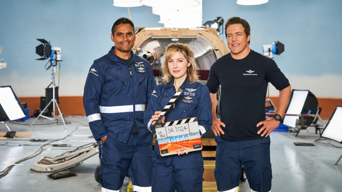 Rob Collins, Emma Hamilton, Stephen Peacocke stand in front of a plane set. Emma holds a clapping board.