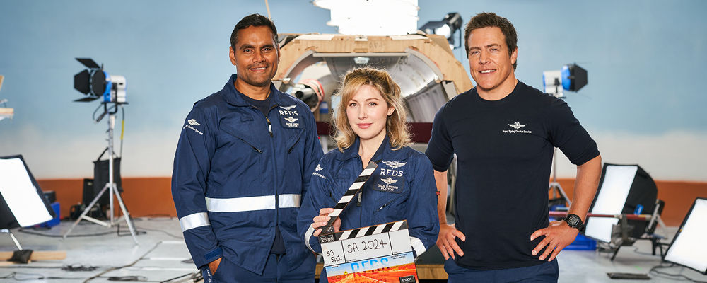 Rob Collins, Emma Hamilton, Stephen Peacocke stand in front of a plane set. Emma holds a clapping board.