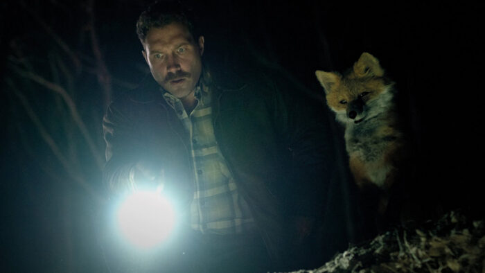 Jai Courtney holds a torch next to a puppet fox.