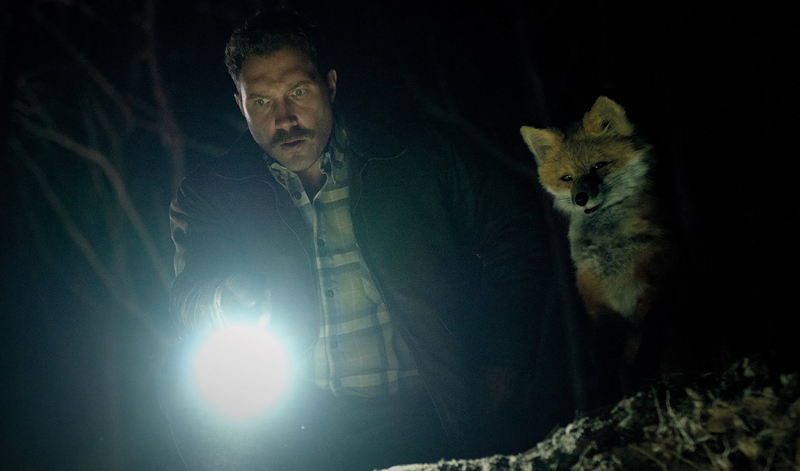 Jai Courtney holds a torch next to a puppet fox.
