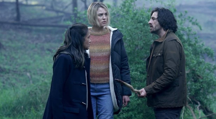 Three people stand in a foggy woodland area. They look deep in conversation.