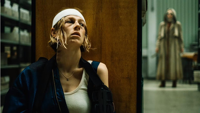 A woman with a bandaged head and beat up appearance hides behind a wall. A menacing figure stands in the distance.