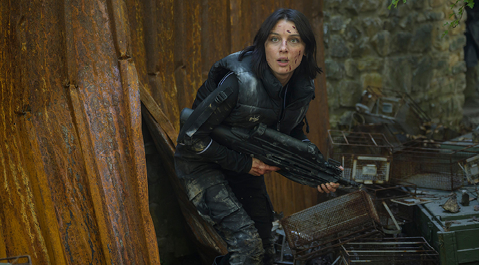 A woman in a black jumpsuit holding a large gun looks around a tree trunk.