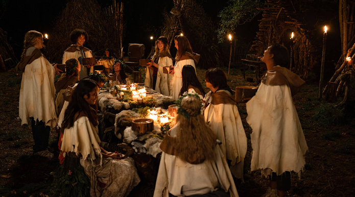 A group of young women sit around a makeshift table at night, lit by candelight. They are all dressed in white and animal furs.