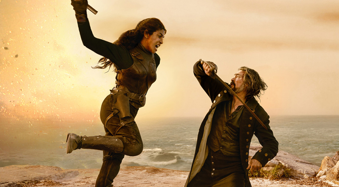 A woman dressed in a black jumpsuit and holding a dagger leaps towards a cowering man.