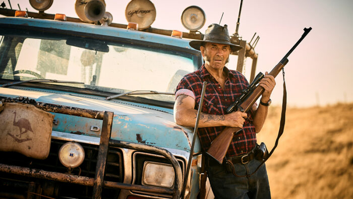 John Jarratt as Mick Taylor leans against a blue ute holding a shotgun.