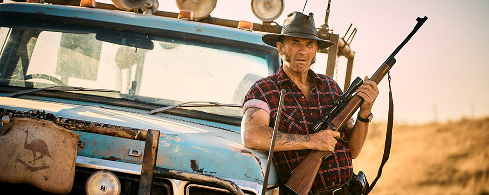 John Jarratt as Mick Taylor leans against a blue ute holding a shotgun.