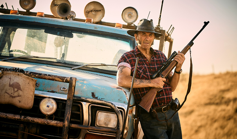 John Jarratt as Mick Taylor leans against a blue ute holding a shotgun.