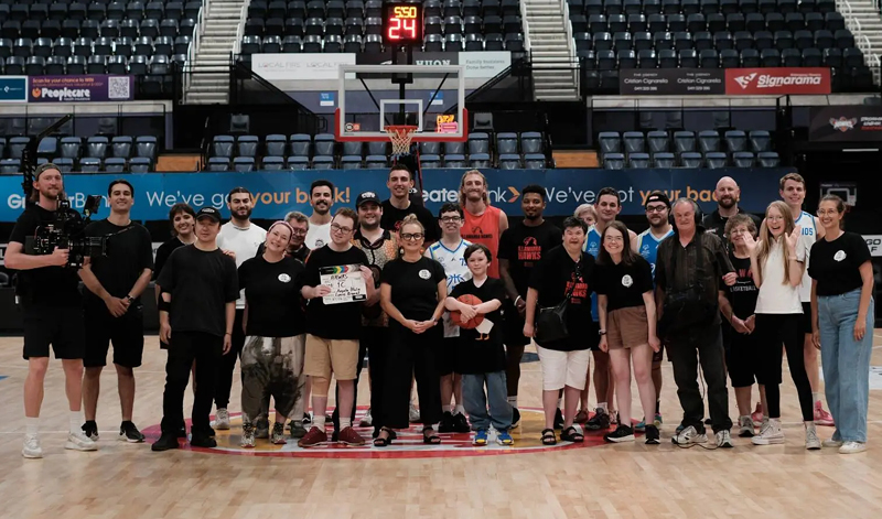 A group of crew members stand in a basketball stadium. A person near the middle holds a clapperboard.