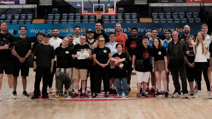 A group of crew members stand in a basketball stadium. A person near the middle holds a clapperboard.