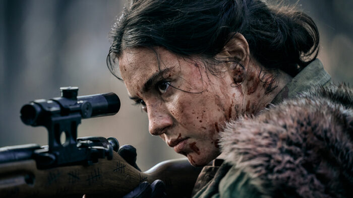A dark haired woman with blood splattered all over her face looks into the scope of a rifle.